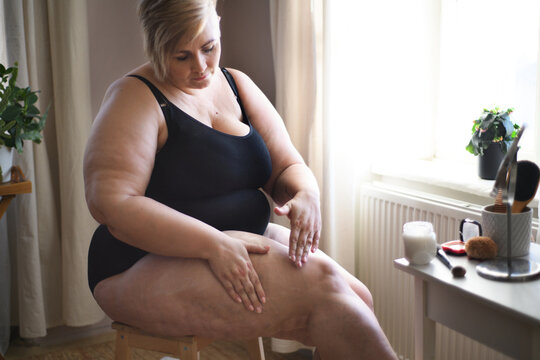 Overweight woman sitting and applying cream on her legs at home, selfcare concept.