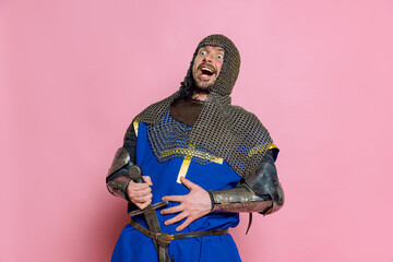 Portrait of man, medieval warrior or knight in special armor with sword laughing isolated over pink...
