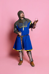 Full-length portrait of man with delightful facial expression in amor with sword, holding glass of vine isolated over pink background