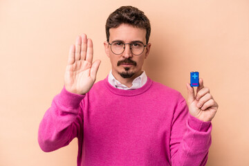 Young caucasian man holding batteries isolated on beige background standing with outstretched hand showing stop sign, preventing you.