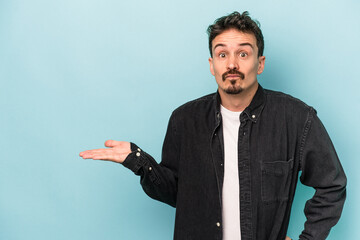 Young caucasian man isolated on blue background showing a copy space on a palm and holding another hand on waist.