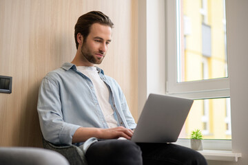 Fototapeta premium Focused determined young bearded businessman or freelancer in casual wear typing on laptop, working on project remotely, researching, writing email to client, web surfing staying at home, side view