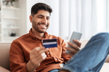 Smiling Arab guy using phone and credit card at home