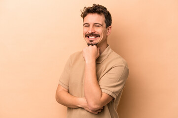 Young caucasian man isolated on beige background smiling happy and confident, touching chin with hand.