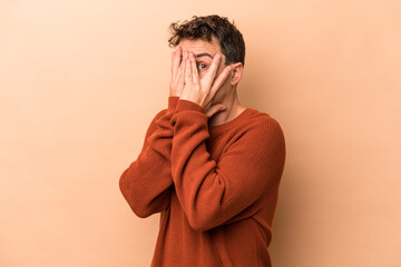 Young caucasian man isolated on beige background blink through fingers frightened and nervous.