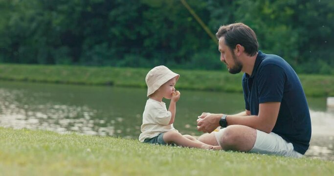 A man sits on a summer afternoon with his young son on the grass near a lake, they watch water sports, eat fruit from a box, the child eagerly reaches for an orange