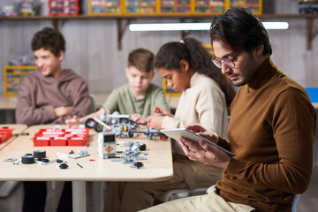 Side view portrait of young male teacher using digital tablet with group of children building robots in background at engineering class, copy space