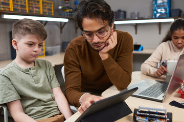 Portrait of young man using digital tablet while teaching robotics class to diverse group of children in modern school