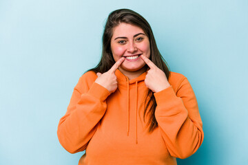 Young caucasian overweight woman isolated on blue background smiles, pointing fingers at mouth.