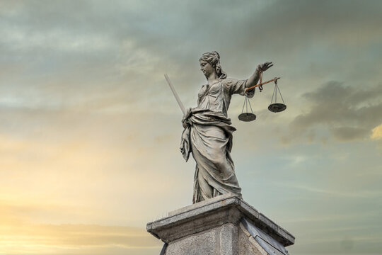Eine Statue der Justitia, die Göttin der Gerechtigkeit, mit Schwert und Waage auf einem Sockel vor einem malerischen Sonnenuntergang