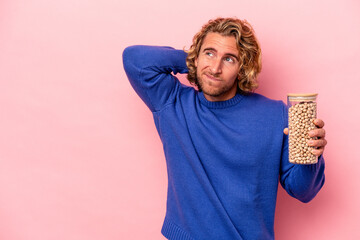 Young caucasian man holding chickpeas isolated on pink background touching back of head, thinking...