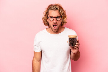 Young caucasian man holding coffee jar isolated on pink background screaming very angry and...