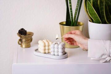Female hand igniting a grey and off-white bubble candles on a concrete tray on white sideboard,...