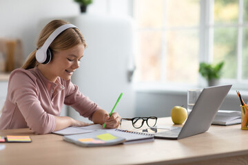 Happy caucasian teen girl blonde in headphones makes notes, study at home at table with laptop in...