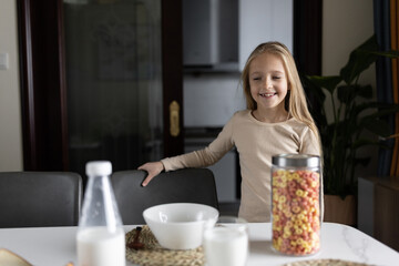 Cute little caucasian girl sitting at table on kitchen early morning and preparing breakfast with colorful cornflakes and milk. Kid enjoying life with healthful food, healthy lifestyle concept