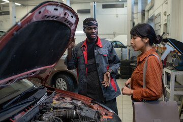 Portrait of black car mechanic talking to female client while standing by open hood in auto repair...