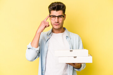 Young caucasian man holding pizzas isolated on yellow background pointing temple with finger, thinking, focused on a task.