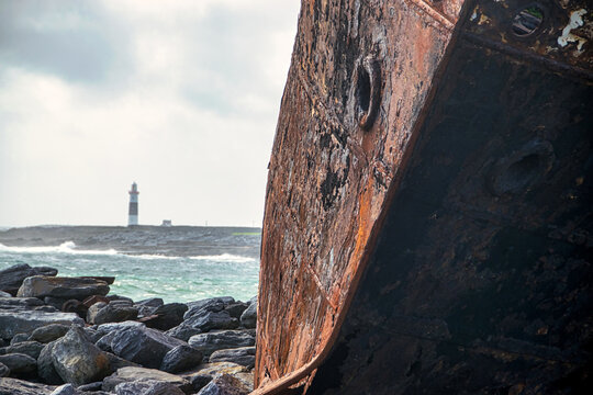 Im Vordergrund erkennt man das verrostete Bug eines Schiffswracks am Steinstrand, im Hintergrund einen Leuchtturm