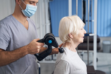 Physiotherapist treating pensioner shoulder with muscle massage gun