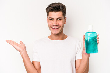 Young caucasian man holding mouthwash isolated on white background showing a copy space on a palm and holding another hand on waist.