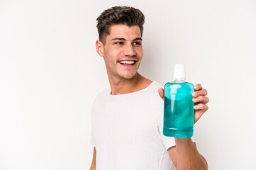 Young caucasian man holding mouthwash isolated on white background looks aside smiling, cheerful and pleasant.
