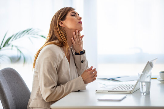 A sick business woman with a sore throat touches the point of pain in her neck and holds a glass of water while sitting in a modern office. The concept of work and disease