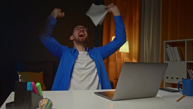 Excited businessman work from home check information at paper report online raise hand yes overjoyed with great sales results. Emotional young male freelancer celebrate success achieve business goal