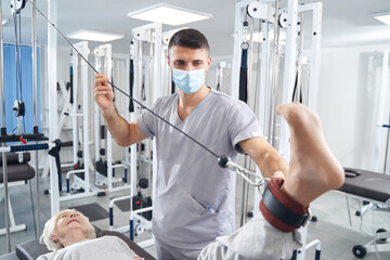 Man medical worker supporting pensioner leg during exercise