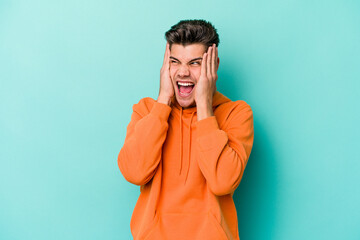 Young caucasian man isolated on blue background covering ears with hands trying not to hear too...