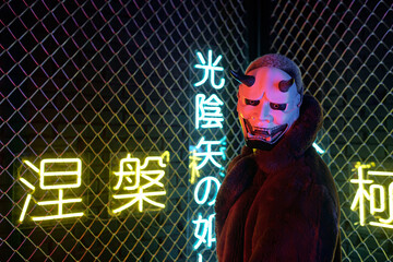 Cyberpunk guy in evil mask standing in front of the bars in darkness with neon hieroglyphs which...