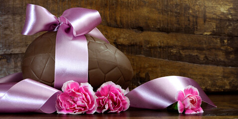 Happy Easter chocolate easter egg with satin pink ribbon and pink flowers against a dark vintage recycled wood timber background with copy space.e. Sized to fit popular social media and web banner.