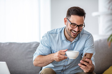 A smiling man makes a video call with friends while sitting on a sofa in his modern home. He uses a smartphone and drinks morning coffee. The concept of happy business people. Blurred background