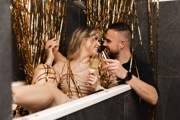 Obraz na płótnie Canvas Portrait of romantic gentle couple woman and man sitting in bathtub drinking champaign, celebrate New Year near lametta