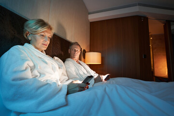 Serious lady and worried man checking news online in bed