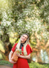 Smiling young woman in a bright red dress walks through a blooming garden. In her hands she holds a wicker shopping bag. She is free and happy. Concept of freedom