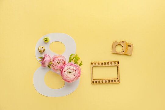 Greeting Card International Women's Day on March 8th. Pink ranunculus decorates the number eight and wooden photo frames on yellow background. Soft focus. Top view.