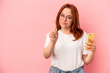 Young caucasian woman holding a mobile phone isolated on pink background showing number one with finger.