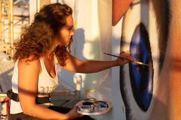 brunette female artist woman in sexy white shirt, in the warm evening sunlight, painting with brush and palette in front of a big mural, paints a peacock butterfly on the wall