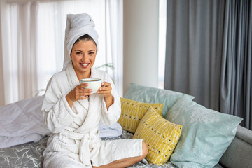 A lazy woman in a bathrobe lies on the bed and enjoys a sunny morning with a cup of aromatic coffee while relaxing in the comfortable bedroom of the hotel room. Easy lifestyle.