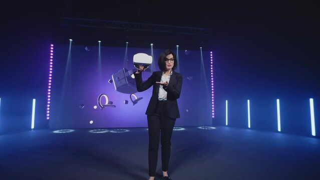 A female speaker in casual clothes in a studio with LED screen holding new virtual reality glasses at a Metaverse presentation and speaking about new modern technologies, in the virtual world