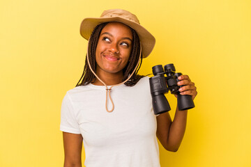 Young african american explorer woman holding a binoculars isolated on yellow background  dreaming of achieving goals and purposes