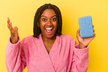 Young african american woman wearing a bathrobe holding a blue sponge isolated on yellow background...