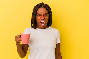 Young african american woman holding a mug isolated on yellow background  screaming very angry and aggressive.