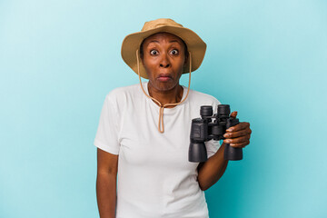 Young african american woman holding binoculars isolated on blue background shrugs shoulders and...
