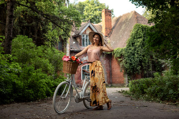 Young caucasian woman in straw hat and summer clothes walk with vintage bicycle with basket with flowers in front of medieval brick mansion in green park. Weekend, leisure activity. Full length.