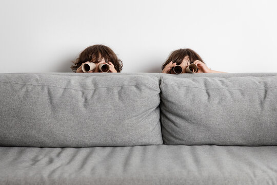 Portrait of twin brothers hiding behind couch looking through paper binoculars