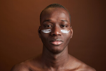 Portrait of African young man applying the cream on his face to protect the skin isolated on brown...