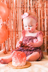 Cute birthday girl celebrating her one year birthday by messily eating a cake surrounded by pink background streamers and balloons