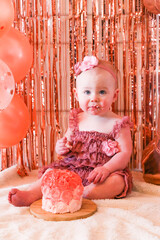 Cute birthday girl celebrating her one year birthday by messily eating a cake surrounded by pink background streamers and balloons