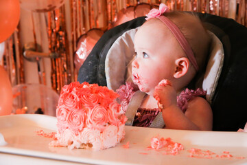 Cute birthday girl celebrating her one year birthday by messily eating a cake surrounded by pink background streamers and balloons